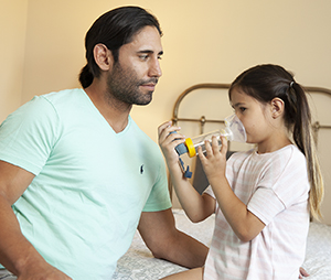 Un hombre ayuda a una niña a usar un inhalador de dosis medida con un espaciador y una máscara.